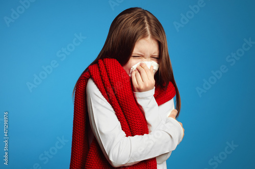 Young kid feels unwell, blows nose in white tissue, suffers from running nose, cold symptoms or allergy, sneezes very often, wearing red scarf