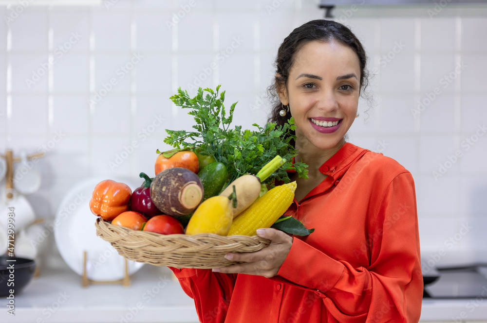 ํYoung woman holding a basket full of fruits and vegetables in the kitchen ํYoung woman holding a basket full of fruits and vegetables in the kitchen