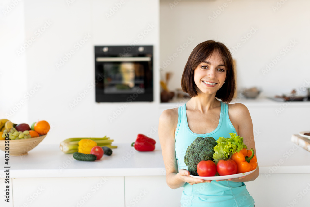 Healthy dinner for slimming and dieting. Happy millennial lady holding plate with vegetables, standing in kitchen