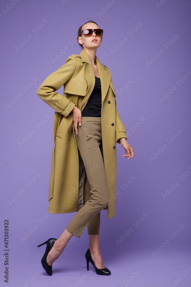 Fashion photo of a beautiful elegant young woman in a pretty yellow mustard coat, pants, trousers posing on a purple lavender background. Studio shot, portrait. Stylish sunglasses Fashion photo of a beautiful elegant young woman in a pretty yellow mustard coat, pants, trousers posing on a purple lavender background. Studio shot, portrait. Stylish sunglasses