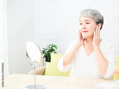 Happy 50s middle aged asian woman touching face skin looking in mirror reflection. Smiling mature old lady pampering  healthy moisturized skin care  aging beauty  skincare treatment cosmetics concept.