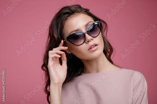 High fashion photo of a beautiful elegant young woman in a pretty pink jumpsuit, sunglasses posing on pink background. Slim figure. Studio Shot. Luxurious hair, shiny curls. Femininity and tenderness