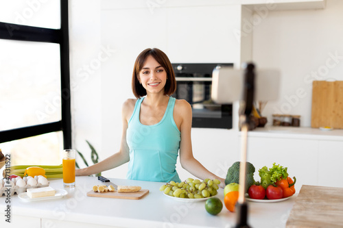 Online lesson of useful cooking. FIt millennial woman making salad  cutting fruits and looking at smartphone in kitchen
