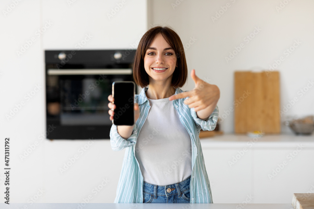 Excited woman holding smartphone with blank screen and pointing on it while standing in kitchen interior, copy space