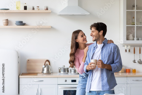 Cheerful millennial european wife hugs husband with cup of hot drink in minimalist kitchen interior