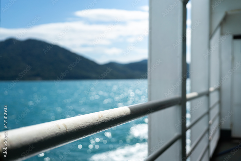 Close-up a white falling prevention rail at corridor walkway of the passenger boat with blurred background of ocean and island view. Transportation and travel scene. Selective focus at railing part. Close-up a white falling prevention rail at corridor walkway of the passenger boat with blurred background of ocean and island view. Transportation and travel scene. Selective focus at railing part.