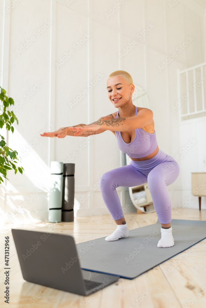 Home workout. Slim young woman in sport uniform doing squat exercises and looking at laptop, watching online lessons Home workout. Slim young woman in sport uniform doing squat exercises and looking at laptop, watching online lessons