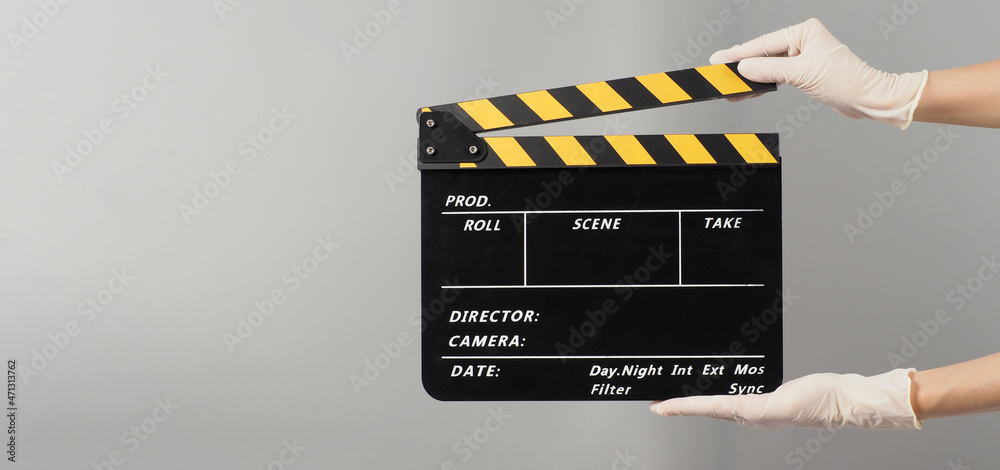 Hand is holding yellow with black clapper board color  and wear medical glove on grey background.