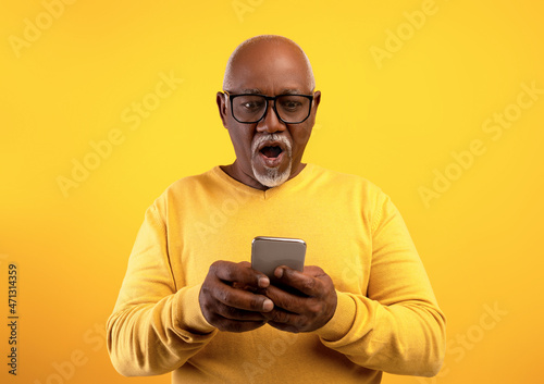 Emotional senior African American man in glasses looking at his smartphone in shock on orange studio background © Prostock-studio