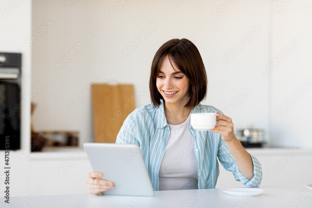 Positive caucasian woman drinking coffee and using digital tablet, surfing internet online, sitting at table in kitchen Positive caucasian woman drinking coffee and using digital tablet, surfing internet online, sitting at table in kitchen