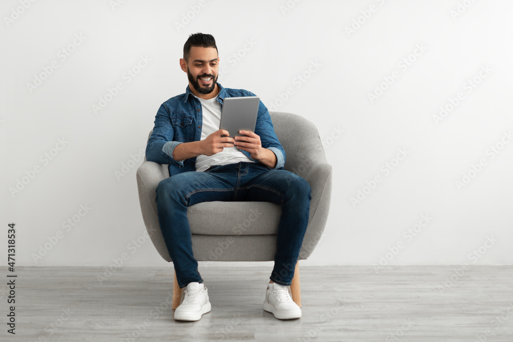 Young Arab man with digital tablet sitting in armchair, working online, having webinar against white studio wall Young Arab man with digital tablet sitting in armchair, working online, having webinar against white studio wall