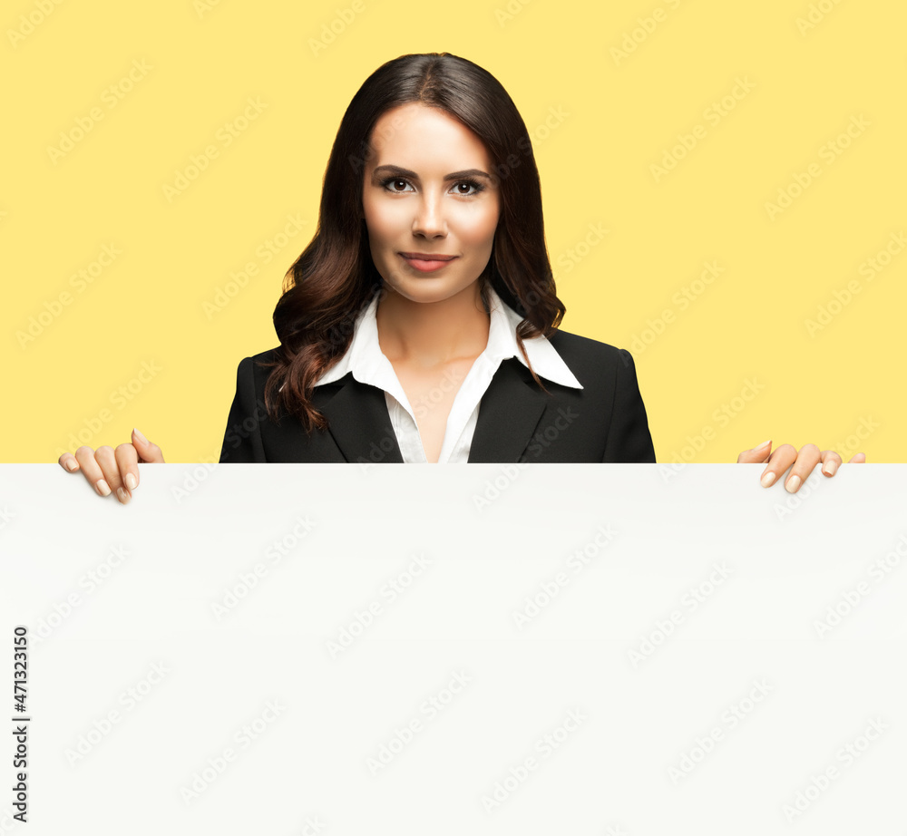 Naklejka premium Happy smiling brunette business woman in confident black suit, standing behind, peeping from blank banner or mock up signboard, isolated over yellow color background.