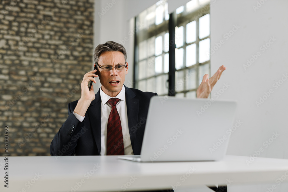 Frustrated businessman communicating with someone over cell phone while working on a computer Frustrated businessman communicating with someone over cell phone while working on a computer