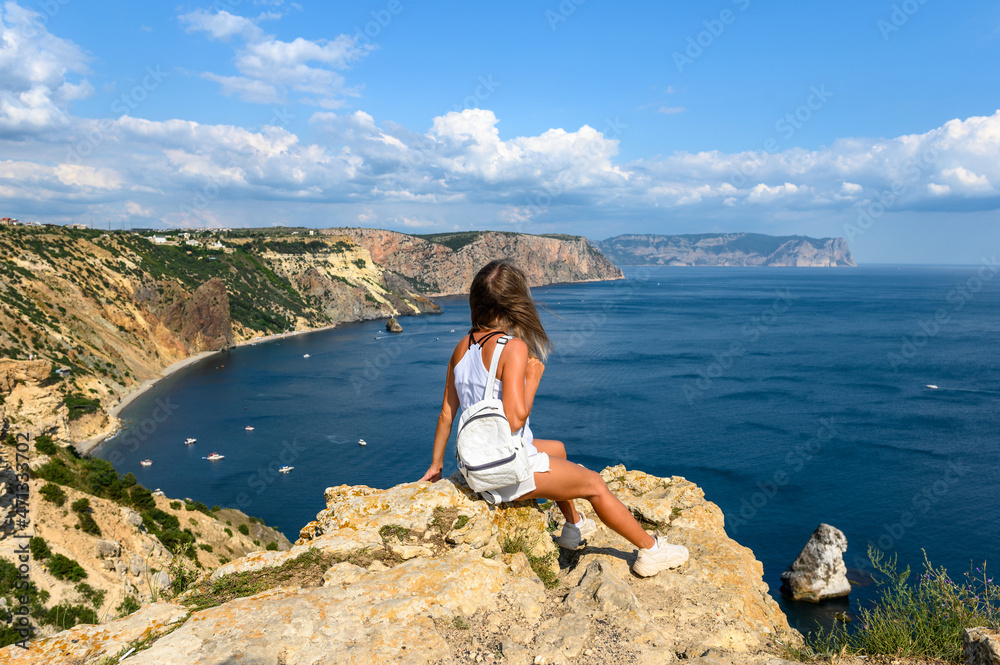 The girl traveler sits on a huge stone on a rock and looks at the sea.