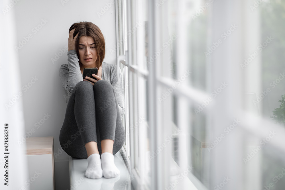 Confused shocked sad depressed european millennial lady sits on windowsill and reading message on smartphone