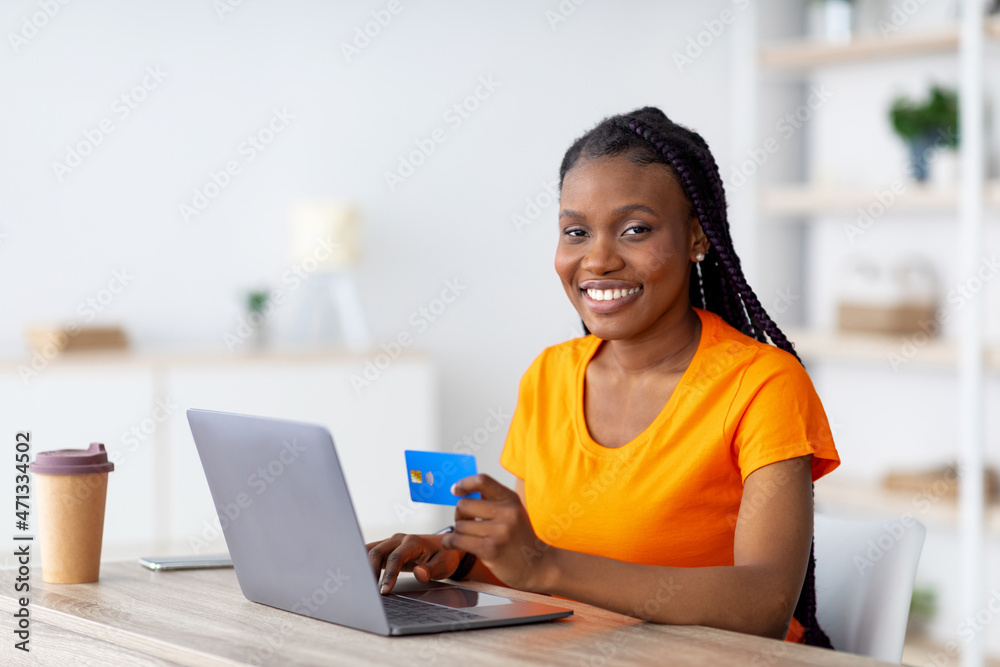Cheerful black woman with laptop and credit card sitting at desk, shopping online, making remote payment at home Cheerful black woman with laptop and credit card sitting at desk, shopping online, making remote payment at home