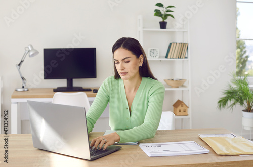 Portrait of young focused woman working with laptop and documents in open space office. Stylish young woman typing on laptop while sitting at desk on background of another workplace. Business concept.