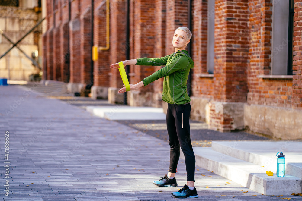 Full height of sportive woman runner workout with fitness resistance band outdoor with old industrial street with red brick facades on background.