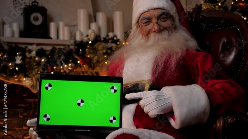 Modern smiling Santa Claus points finger at laptop green screen markers. Positive Santa Claus shows hand on computer mock up chroma key display, Christmas online shopping, New Year gifts, holidays