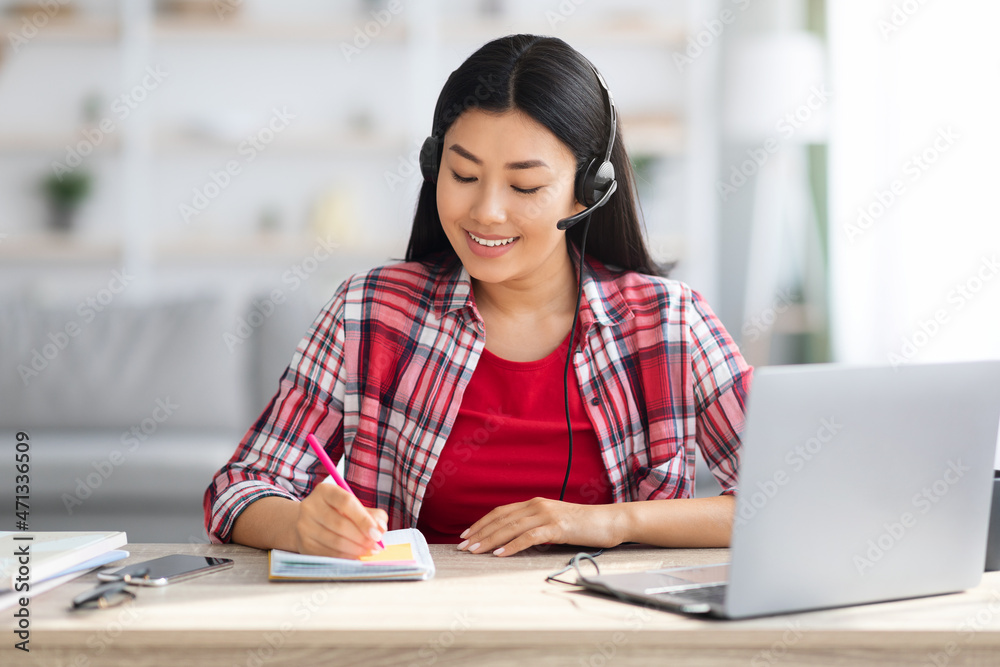 Asian Female In Headset Taking Notes While Study With Laptop At Home Asian Female In Headset Taking Notes While Study With Laptop At Home