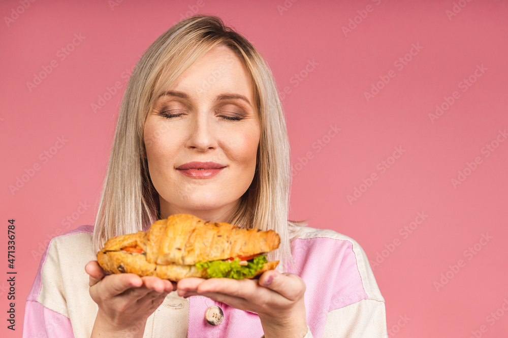 Portrait of young beautiful hungry woman eating croissant sandwich. Isolated portrait of woman with fast food over pink background. Diet concept.