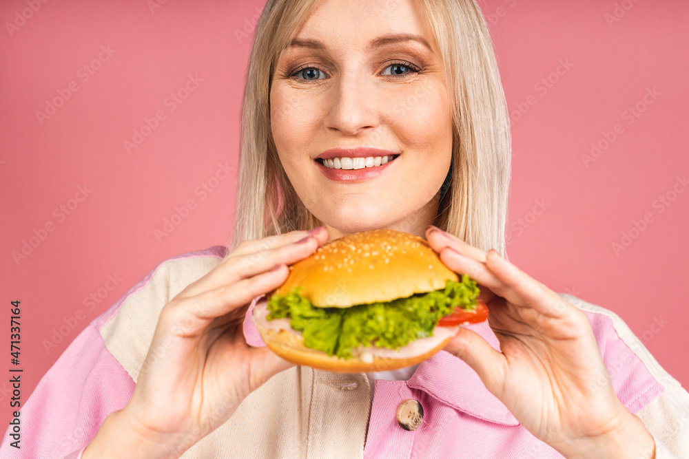 Cheerful young hungry beautiful blonde woman holding cheeseburger or burger holding in hands isolated over pink background. Junk food concept. Cheerful young hungry beautiful blonde woman holding cheeseburger or burger holding in hands isolated over pink background. Junk food concept.