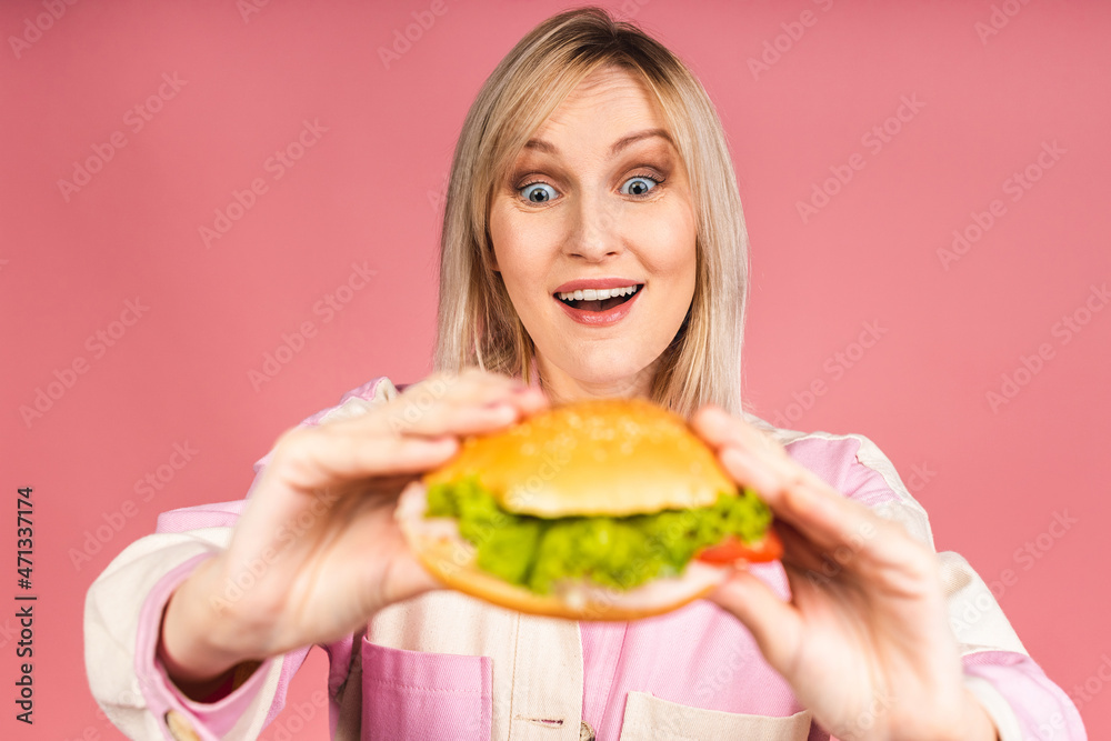 Cheerful young hungry beautiful blonde woman holding cheeseburger or burger holding in hands isolated over pink background. Junk food concept. Cheerful young hungry beautiful blonde woman holding cheeseburger or burger holding in hands isolated over pink background. Junk food concept.