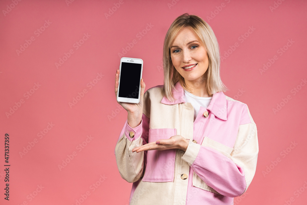 Naklejka premium Portrait of attractive young blonde woman with white smile, showing empty smartphone screen, demonstrate an app or online shopping store, isolated over pink background.
