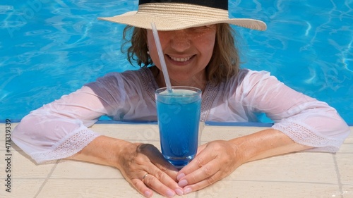 happy senior woman relaxing near blue outdoor swimming pool with blue cocktail wearing straw hat. People are enjoying their summer vacation. All inclusive