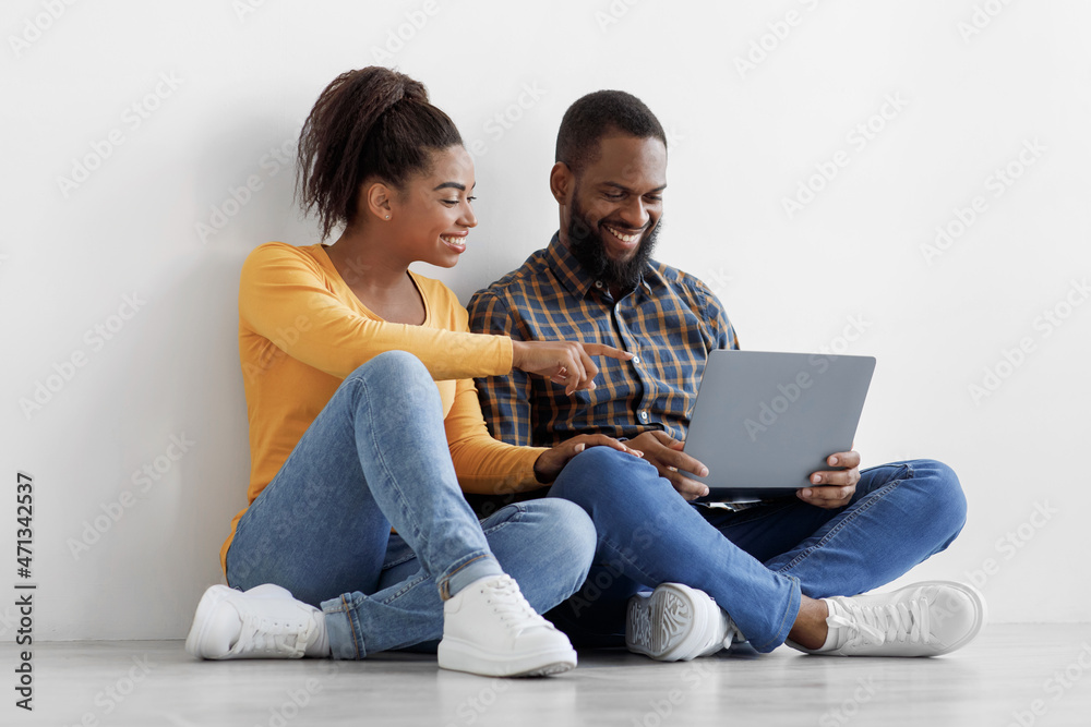 Happy young african american guy and lady look in laptop, choosing interior, sit on floor