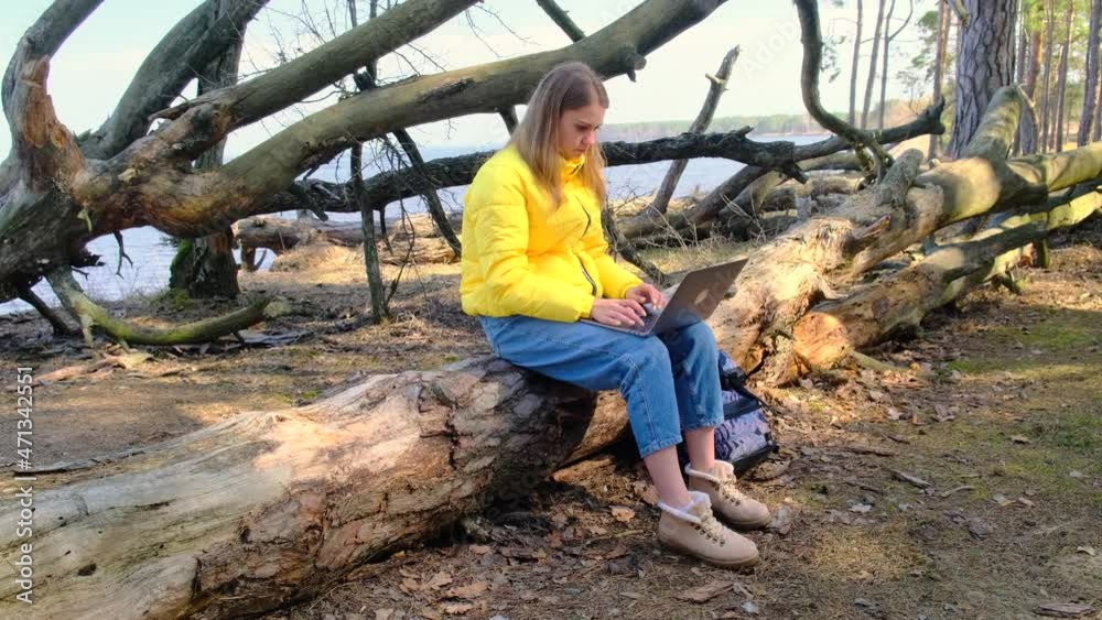 Downshifting or freelancing. A young woman uses a laptop sitting on a log or fallen tree in the woods against the backdrop of a river on a sunny day. Girl wears a yellow jacket. jeans and boots. 4k Downshifting or freelancing. A young woman uses a laptop sitting on a log or fallen tree in the woods against the backdrop of a river on a sunny day. Girl wears a yellow jacket. jeans and boots. 4k