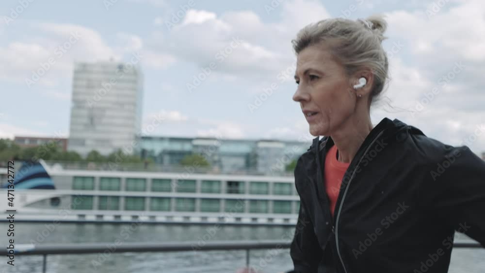 Mature woman in earphones running on street near city river Mature woman in earphones running on street near city river
