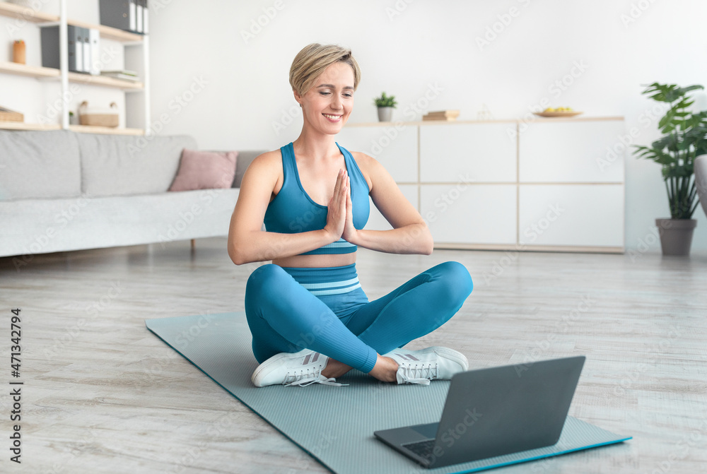 Fototapeta premium Woman meditating sitting in lotus position on mat with laptop