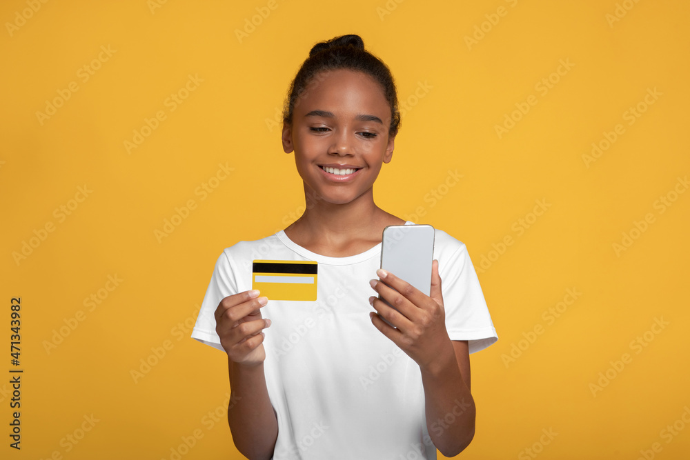 Glad teenage black girl pupil in white t-shirt hold credit card and paying with smartphone Glad teenage black girl pupil in white t-shirt hold credit card and paying with smartphone