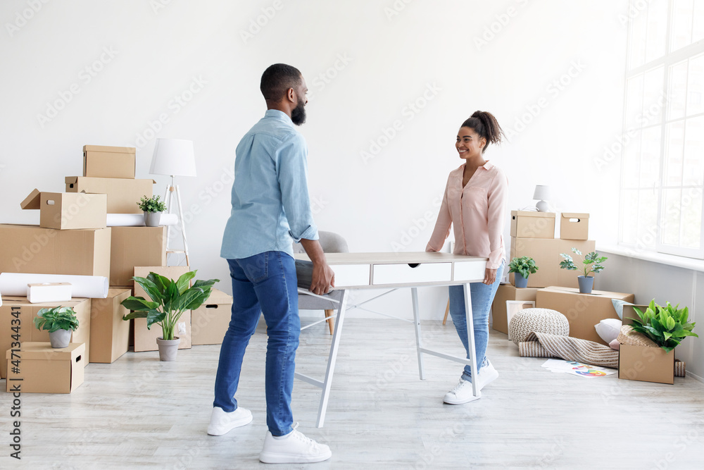 Happy young african american husband and wife carry table to new house with cardboard boxes