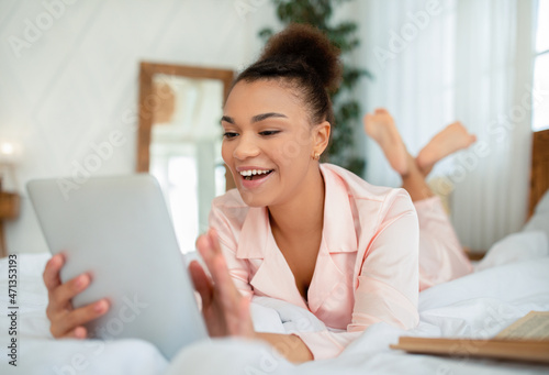 Smiling african american lady in pajama laying on bed and using modern digital tablet, copy space