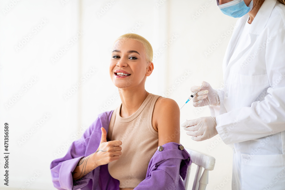 Fototapeta premium Happy millennial lady approving of covid-19 vaccination, gesturing thumbs up before coronavirus vaccine injection