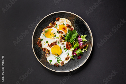 plate of fried eggs with dried tomatoes and fresh salad leaves on dark background, top view
