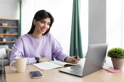 Woman sitting at desk, using computer and writing in notebook
