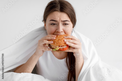 Disappointed hungry caucasian millennial female eating big burger sitting on white bed in bedroom
