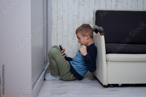 mały chłopak siedzi między fotelem a szafą, trzyma w dłoni telefon jest zdenerwowany © ftomasz