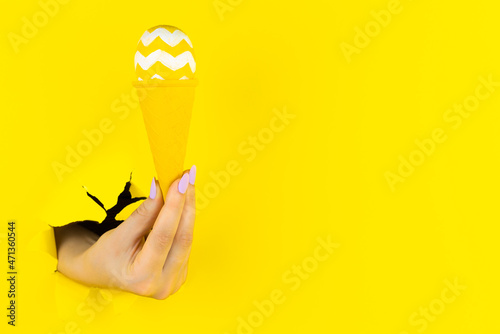 Abstract yellow background with female hand is holding decorated egg in colorful ice cream cone. Minimal art design. Minimalist meal, fashion style. Festive food court. Happy Easter festival concept.