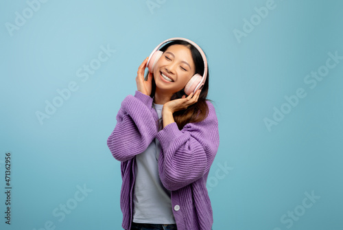 Excited asian woman listening to music with closed eyes using wireless headphones, standing over blue background