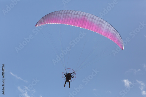 A man flying a pink and white motorized parasail moves away from the camera through a blue sky with a few bits of puffy white cloud above him.