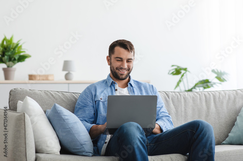 Smiling mature caucasian guy with beard sitting on sofa look at laptop in living room interior at spare time