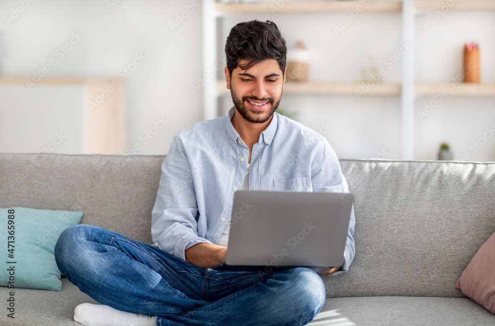 Portrait of arab man sitting on sofa, using laptop indoors in living room at home, free space