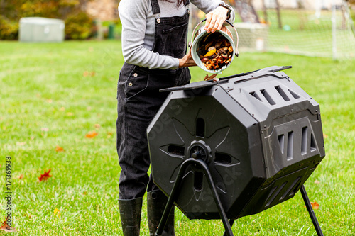 A woman is dumping a small bin of kitchen scraps into an outdoor tumbling composter in backyard garden. These plastic units with metal legs can turn around for better aeration and quick composting.