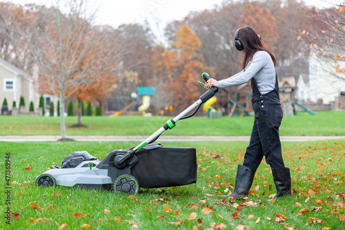 a young caucasian woman wearing overalls and noise blockers is operating a lawnmover on grass in late autumn. There are fallen leaves and a suburban neighborhood setting in background. photo