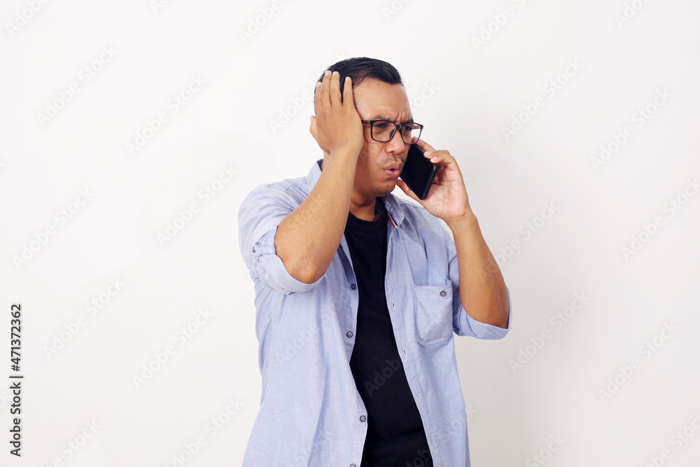 Young Asian man using smartphone stressed with hand on head, shocked with shame and surprise face, angry and frustrated over white wall background. Fear and upset for mistake. Young Asian man using smartphone stressed with hand on head, shocked with shame and surprise face, angry and frustrated over white wall background. Fear and upset for mistake.