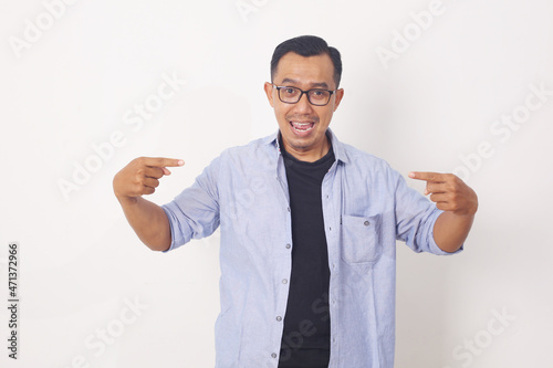 Portrait of funny young Asian man smiling and pointing to presenting something in front his body. Isolated on white background with copyspace